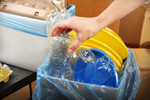 A close-up of a person's hand holding a clear plastic bottle, which is partially filled with water, poised above a large blue plastic bin lined with a transparent plastic bag. Inside the bin, there are multiple yellow and blue plastic buckets, stacked or leaning against each other. The background reveals a wooden surface and part of an indoor storage or office space, suggesting an organised area for rubbish collection or disposal. The scene highlights the handling of recyclable materials, consistent with rubbish removal services offered by companies such as Office Clearance Willesden, serving the Willesden area in northwest London. Lighting is natural and even, emphasizing the textures of the plastic containers, the smooth surface of the bottle, and the glossy finish of the plastic lining, all relevant to waste sorting and disposal activities.