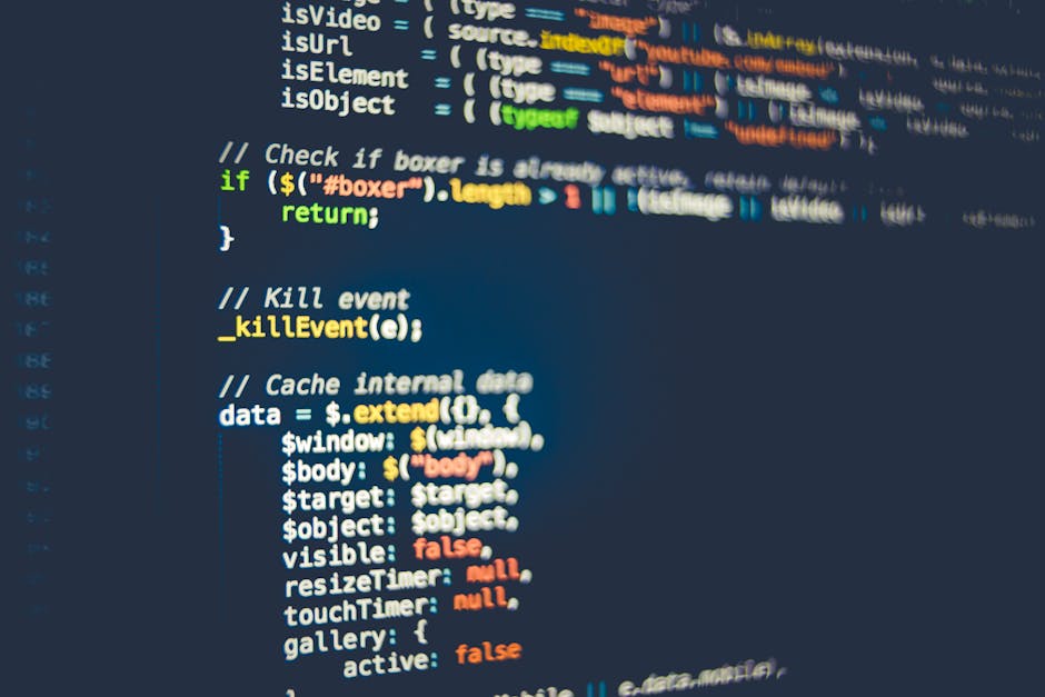 A silver laptop with a black keyboard is positioned on a light wooden desk, displaying lines of code on its screen with a dark background and multicolored text. Next to the laptop, there is a closed w