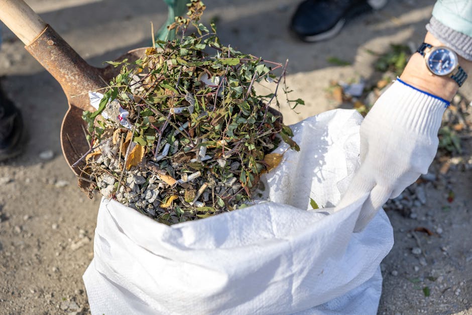 A person wearing light blue jeans and white sneakers is crouching on asphalt pavement during daytime, holding a small collection of discarded debris including a white plastic container, a fragment of 
